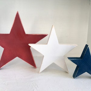 Puede incluir: Tres estrellas de madera en rojo, blanco y azul. Las estrellas est&aacute;n pintadas en un estilo desgastado y est&aacute;n dispuestas en orden de tama&ntilde;o, con la estrella m&aacute;s grande en la parte posterior y la estrella m&aacute;s peque&ntilde;a en la parte delantera.