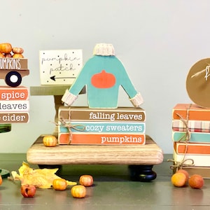 May include: A fall-themed display with three stacks of wooden books. Each stack features a different fall-themed design, including a truck with pumpkins, a sweater with a pumpkin, and a pumpkin with the text "hello fall".