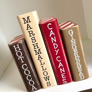 K&ouml;nnte beinhalten: Vier Holzbl&ouml;cke in verschiedenen Farben bemalt, jeder mit einer festlichen Leckerei beschriftet. Die Bl&ouml;cke sind braun, beige, rot und beige, mit den W&ouml;rtern "Hot Cocoa", "Marshmallows", "Candy Canes" und "Gingerbread" in wei&szlig;er Schrift. Jeder Block hat eine gestreifte Oberseite.