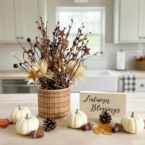 May include: A cylindrical container with a brown and yellow plaid pattern holds a decorative arrangement of twigs, dark berries, cream-colored flowers, and brown stars. A rectangular sign reads Autumn Blessings, surrounded by white pumpkins and pinecones.
