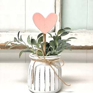 May include: A white ceramic planter with a ribbed texture and a brown twine bow. The planter is filled with green leafy plants and a pink wooden heart on a stick.