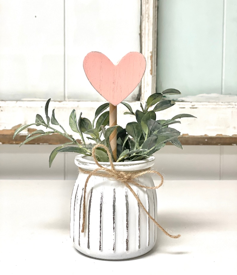 May include: A white ceramic planter with a ribbed texture and a brown twine bow. The planter is filled with green leafy plants and a pink wooden heart on a stick.