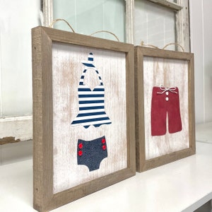 May include: Two framed wood signs with a white background. The sign on the left features a blue and white striped swimsuit with red buttons. The sign on the right features red swim trunks with a white bow and a white button.