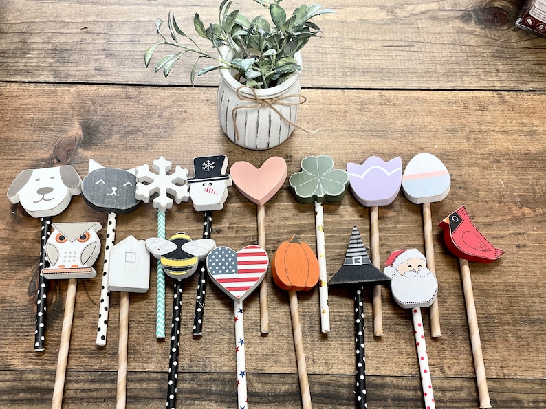 May include: A set of 15 wooden painted ornaments on sticks. The ornaments feature various designs, including a snowman, a heart, a bee, a pumpkin, a cardinal, a house, a snowflake, a cat, a dog, a shamrock, a tulip, an egg, a witch's hat, a Santa Claus, and an American flag.