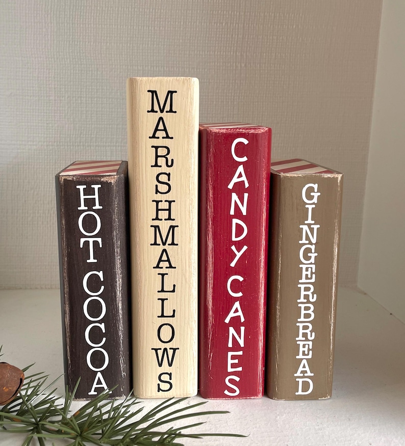 K&ouml;nnte beinhalten: Vier Holzbl&ouml;cke mit den W&ouml;rtern "Hei&szlig;e Schokolade", "Marshmallows", "Zuckerstangen" und "Lebkuchen". Die Bl&ouml;cke sind in Braun, Creme, Rot und Beige bemalt. Jeder Block hat wei&szlig;en Text, einige haben gestreifte Akzente.