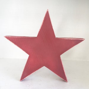 Puede incluir: Una gran estrella de cinco puntas de madera, pintada en un tono rosa polvoriento profundo. La estrella tiene un acabado ligeramente desgastado, lo que le da un aspecto vintage.