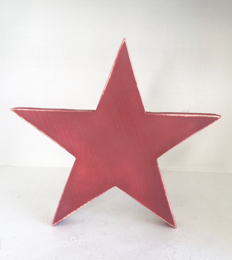 Puede incluir: Una gran estrella de cinco puntas de madera, pintada en un tono rosa polvoriento profundo. La estrella tiene un acabado ligeramente desgastado, lo que le da un aspecto vintage.