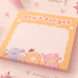 May include: A square, orange sticky note pad with the words "don't forget" in orange lettering. The pad features cartoon characters and a blank space for notes. The words "Amnesia Trio" are written below.
