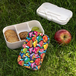 May include: A white lunchbox with three compartments, filled with biscuits and grapes. The lunchbox has a colourful mushroom design on the lid.