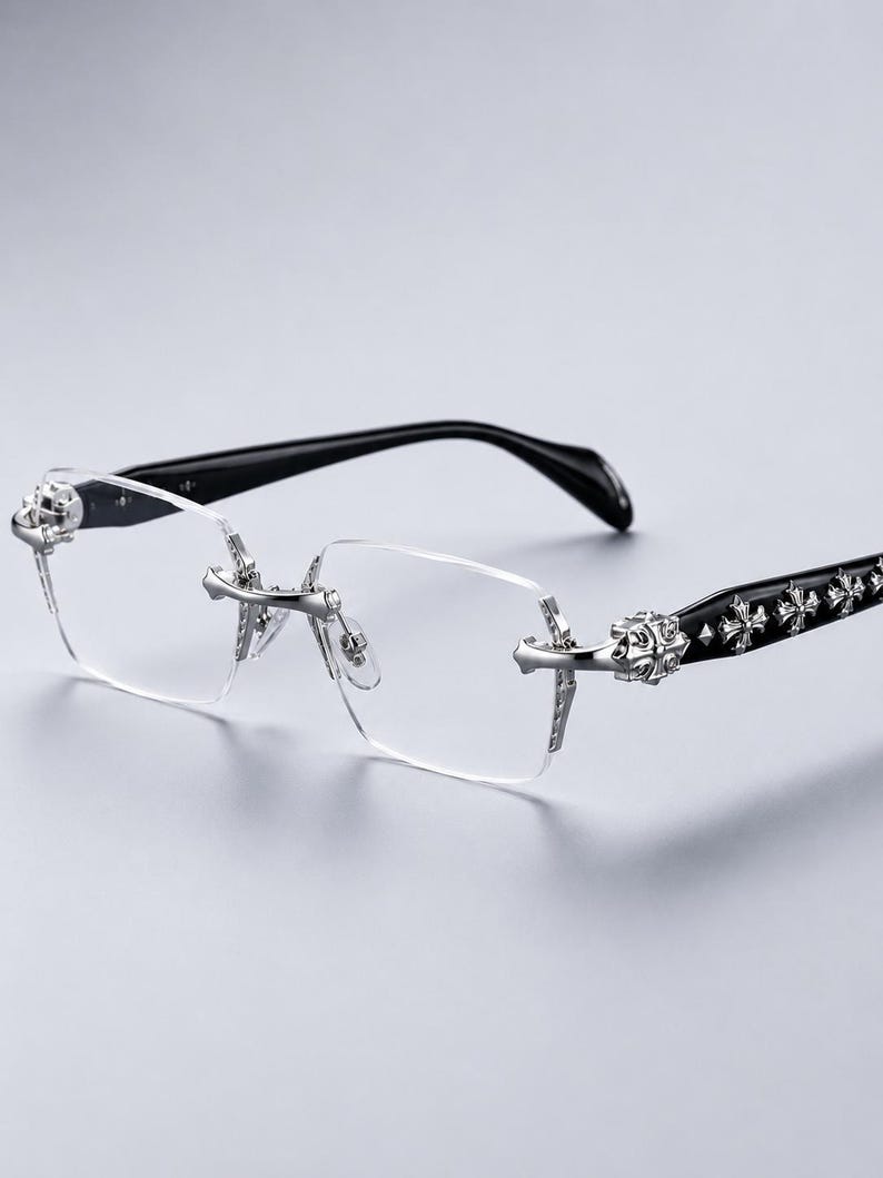K&ouml;nnte beinhalten: Randlose Brille mit klaren, rechteckigen Gl&auml;sern und silberfarbenen Metallbeschl&auml;gen an Steg und Scharnieren. Die B&uuml;gel sind schwarz und mit einer Reihe von erhabenen, silberfarbenen Kreuzemblemen an den Au&szlig;enseiten versehen.