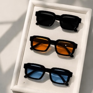 Peut inclure: Trois paires de lunettes de soleil rectangulaires à monture noire épaisse disposées verticalement. La paire du haut a des verres noirs, la paire du milieu a des verres orange vif et la paire du bas a des verres bleu clair, alignées en rangée.