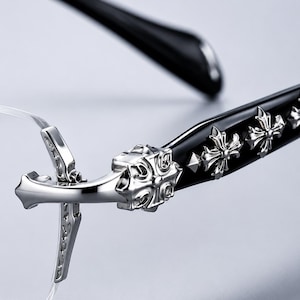 K&ouml;nnte beinhalten: Randlose Brille mit schwarzen B&uuml;geln, die mit silberfarbenen Kreuzemblemen aus Metall verziert sind. Der Scharnierbereich zeigt ein detailliertes silberfarbenes Kreuzdesign, das mit der Glasfassung verbunden ist und ein dekoratives Element bildet.