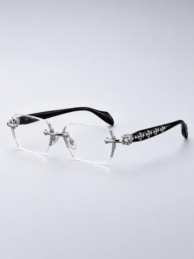 K&ouml;nnte beinhalten: Randlose Brille mit klaren, rechteckigen Gl&auml;sern und silberfarbenen Metallbeschl&auml;gen an Steg und Scharnieren. Die B&uuml;gel sind schwarz und mit einer Reihe dekorativer, silberfarbener Kreuz-Embleme an den Au&szlig;enseiten versehen.
