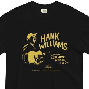 Puede incluir: Camiseta negra con una imagen amarilla de Hank Williams tocando una guitarra. El texto "I heard that Lonesome Whistle Blow" está impreso debajo de la imagen. El texto "No. K11054-A" está impreso en la esquina inferior izquierda.