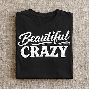 May include: Black t-shirt with the white text "Beautiful CRAZY" in a cursive and block font. The shirt is folded neatly on a neutral-colored surface. The text is the focal point of the image.
