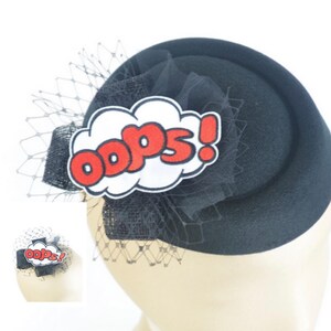 May include: Black pillbox hat with a comic book style "Oops!" speech bubble. The hat features a black bow, netting, and a red and white graphic. A stylish accessory for themed events.