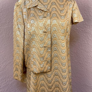 May include: A vintage two-piece suit in a light peach color with a repeating wave pattern in gold and silver. The jacket has short sleeves and a collar, with three buttons down the front. The skirt is knee-length.