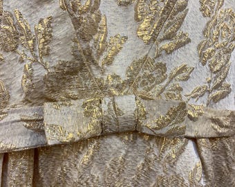 May include: Close-up of a formal dress with a gold floral pattern on a cream background. The dress features a bow at the waist. The fabric has a textured appearance, and the gold detailing adds a touch of elegance.