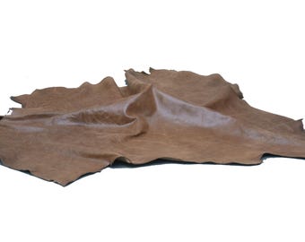 May include: A single, irregular piece of medium brown leather hide with a smooth, matte finish. The material features natural grain variations and folds, displaying an organic, raw edge shape that is characteristic of a full, uncut animal hide.