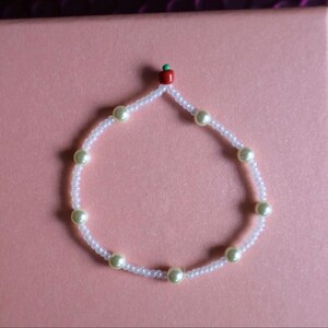 May include: A delicate bracelet featuring white seed beads and larger, pale green pearls. A small red and green apple charm adorns the top of the bracelet. The bracelet is displayed on a pink surface.
