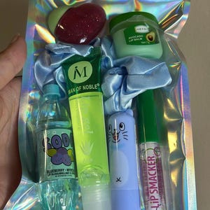 May include: A holographic pouch containing a heart-shaped red glitter balm, a green avocado lip balm, a light blue scrunchie, a blueberry soda-shaped lip gloss, a green Man of Noble aloe lip gel, a light blue cat-themed balm, and a Lip Smacker gloss.
