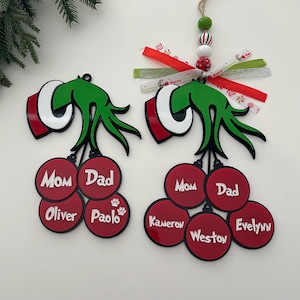 May include: Two green Grinch-themed Christmas ornaments featuring red baubles with white text. One ornament has names Mom, Dad, Oliver, and Paolo with a paw print. The other has Mom, Dad, Kameron, Weston, and Evelynn, topped with ribbons and beads.