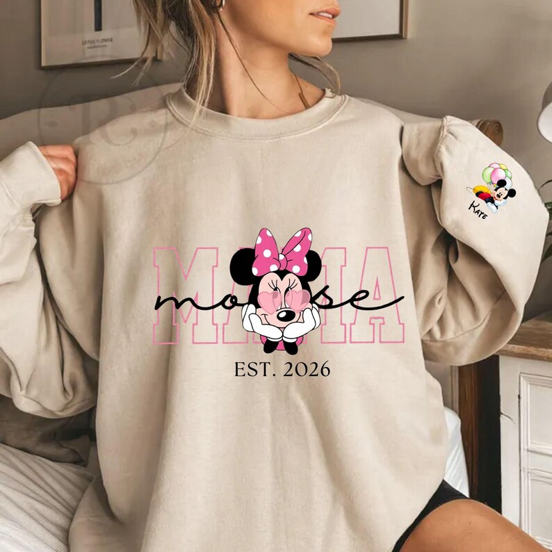 May include: Beige sweatshirt with a Minnie Mouse graphic, pink bow, and the text "Mama Mouse" in pink. The sweatshirt also has "EST. 2026". A small Minnie Mouse graphic with balloons and the name "Kate" is on the sleeve.
