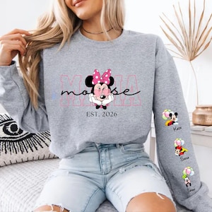 May include: A grey sweatshirt with a Minnie Mouse graphic and the text "MAMA mouse EST. 2026". The sleeve features Mickey Mouse, Minnie Mouse, and Goofy graphics with names. The design incorporates pink and black detailing.