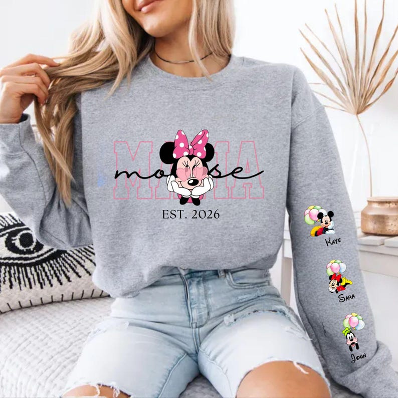 May include: A grey sweatshirt with a Minnie Mouse graphic and the text "MAMA mouse EST. 2026". The sleeve features Mickey Mouse, Minnie Mouse, and Goofy graphics with names. The design incorporates pink and black detailing.