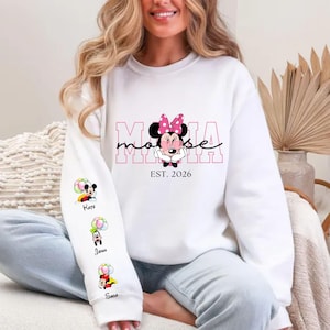May include: White sweatshirt with a Minnie Mouse graphic, pink bow, and the word "Mama" in pink. The sleeve displays cartoon characters with names. The text "EST. 2026" is below the Minnie Mouse graphic. A casual, long-sleeved top.