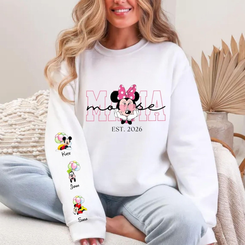 May include: White sweatshirt with a Minnie Mouse graphic, pink bow, and the word "Mama" in pink. The sleeve displays cartoon characters with names. The text "EST. 2026" is below the Minnie Mouse graphic. A casual, long-sleeved top.