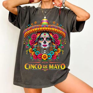 Op de afbeelding: Donkergrijs t-shirt met een kleurrijk ontwerp voor Cinco de Mayo. Het ontwerp bevat een suikerschedel met een sombrero, omringd door bloemen. De tekst "CINCO DE MAYO" is in goud weergegeven.
