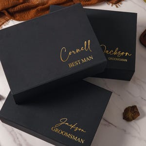 May include: Three black gift boxes with gold lettering. The top box reads "Cornell BEST MAN", the middle box reads "Jackson GROOMSMAN", and the bottom box reads "Jackson GROOMSMAN". The boxes are arranged on a marble surface.