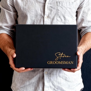 Peut inclure: Une bo&icirc;te cadeau rectangulaire noire avec le pr&eacute;nom Steven &eacute;crit en lettres cursives dor&eacute;es au-dessus du mot GROOMSMAN en lettres dor&eacute;es sans empattement. La bo&icirc;te pr&eacute;sente une finition mate lisse et est tenue horizontalement.