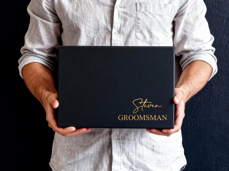 Peut inclure: Une bo&icirc;te cadeau rectangulaire noire avec le pr&eacute;nom Steven &eacute;crit en lettres cursives dor&eacute;es au-dessus du mot GROOMSMAN en lettres dor&eacute;es sans empattement. La bo&icirc;te pr&eacute;sente une finition mate lisse et est tenue horizontalement.