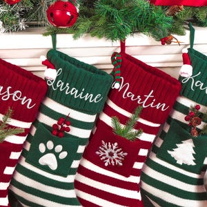 Op de afbeelding: Vier gepersonaliseerde kerstsokken in rood en groen, elk met een naam in wit geborduurd. De sokken hebben witte strepen en decoratieve elementen zoals een pootafdruk, een sneeuwvlok en een kerstboom. Groen en ornamenten maken de feestelijke scène compleet.