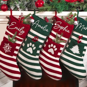 Peut inclure: Quatre bas de Noël rayés rouges et verts, chacun avec un nom brodé en blanc. Un bas a un flocon de neige, deux ont des empreintes de pattes et un a un sapin de Noël. Des décorations et des ornements sont visibles en arrière-plan.