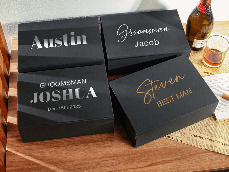 Peut inclure: Quatre bo&icirc;tes cadeaux rectangulaires noires avec du texte personnalis&eacute;. Une bo&icirc;te indique Austin, une autre Groomsman Jacob, la troisi&egrave;me GROOMSMAN JOSHUA Dec 15th.2025, et la quatri&egrave;me Steven BEST MAN. Le texte est en finitions m&eacute;talliques argent et or.