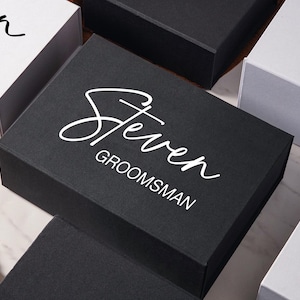 Peut inclure: Une bo&icirc;te cadeau rectangulaire noire avec le pr&eacute;nom Steven imprim&eacute; en blanc et le mot GROOMSMAN dans une police sans empattement. La bo&icirc;te est entour&eacute;e d'autres bo&icirc;tes cadeaux blanches et noires avec un texte similaire sur une surface en marbre.