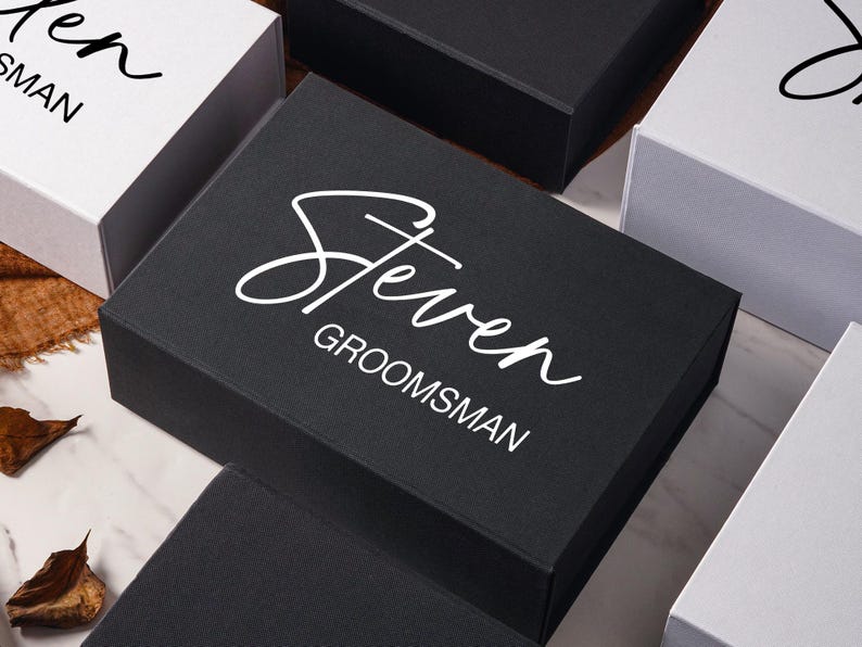 Peut inclure: Une bo&icirc;te cadeau rectangulaire noire avec le pr&eacute;nom Steven imprim&eacute; en blanc et le mot GROOMSMAN dans une police sans empattement. La bo&icirc;te est entour&eacute;e d'autres bo&icirc;tes cadeaux blanches et noires avec un texte similaire sur une surface en marbre.