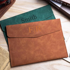 May include: Two rectangular document folders with envelope-style flaps. One folder is dark green with the name Smith engraved on the front. The other folder is brown with the initials FR engraved on the front. Both feature a smooth matte finish.