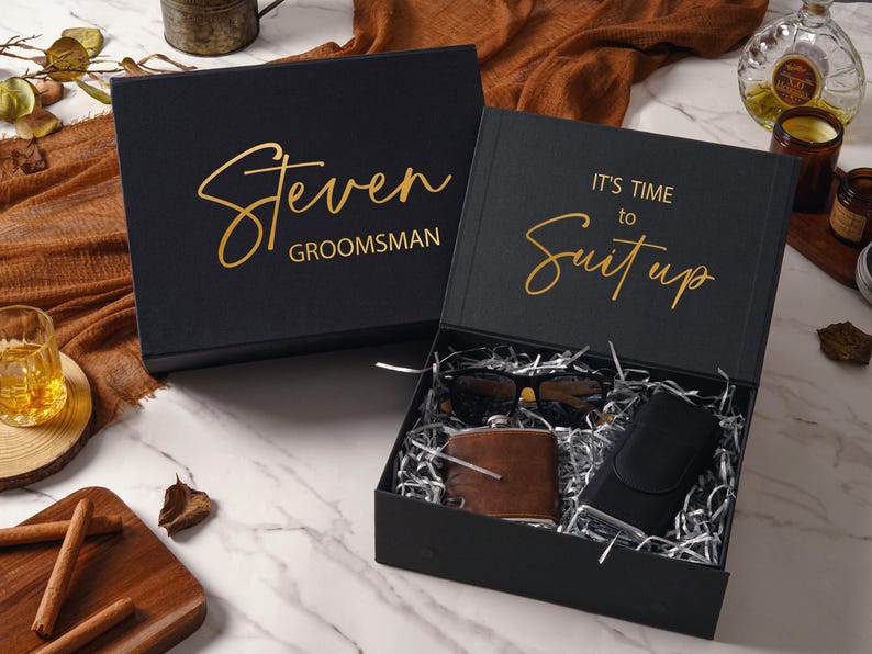 Peut inclure: Une bo&icirc;te cadeau noire avec du texte dor&eacute; indiquant Steven GROOMSMAN et IT'S TIME to Suit up. &Agrave; l'int&eacute;rieur, du papier m&eacute;tallis&eacute; argent&eacute; entoure une flasque gain&eacute;e de cuir marron, un &eacute;tui &agrave; lunettes noir et des lunettes de soleil noires.