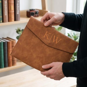 May include: A tan leather document folder with a flap closure featuring the embossed letters K|W on the front. The folder has a smooth, matte texture and a rectangular shape with rounded corners, designed to hold papers or a slim electronic device.