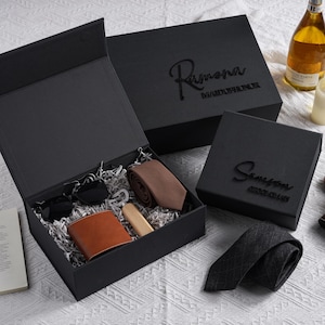 Pode incluir: Um conjunto de presente preto contendo uma petaca de couro castanho, uma gravata castanha, um abre-garrafas de madeira e óculos de sol pretos sobre papel prateado picado. Uma segunda caixa preta apresenta o texto em relevo Ramona Maid of Honor.
