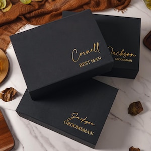 Peut inclure: Trois bo&icirc;tes cadeaux rectangulaires noires avec des inscriptions en feuille d'or. Une bo&icirc;te porte le nom Cornell et le titre BEST MAN, tandis que deux autres portent le nom Jackson et le titre GROOMSMAN en police cursive et sans-serif.