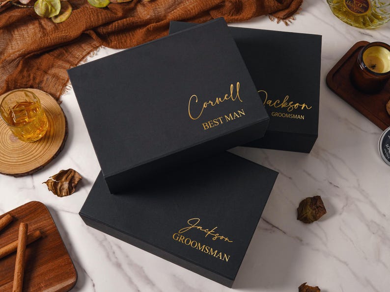 Peut inclure: Trois bo&icirc;tes cadeaux rectangulaires noires avec des inscriptions en feuille d'or. Une bo&icirc;te porte le nom Cornell et le titre BEST MAN, tandis que deux autres portent le nom Jackson et le titre GROOMSMAN en police cursive et sans-serif.