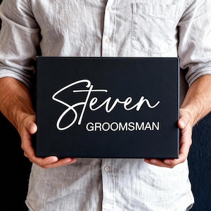 Peut inclure: Une bo&icirc;te cadeau rectangulaire noire portant le pr&eacute;nom Steven &eacute;crit en lettres cursives blanches, avec le mot GROOMSMAN imprim&eacute; en lettres blanches sans empattement juste en dessous du pr&eacute;nom sur le devant de la bo&icirc;te.