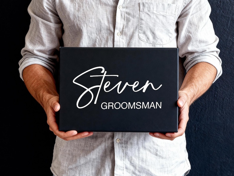 Peut inclure: Une bo&icirc;te cadeau rectangulaire noire portant le pr&eacute;nom Steven &eacute;crit en lettres cursives blanches, avec le mot GROOMSMAN imprim&eacute; en lettres blanches sans empattement juste en dessous du pr&eacute;nom sur le devant de la bo&icirc;te.
