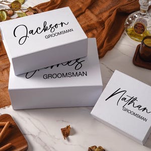 Peut inclure: Trois bo&icirc;tes cadeaux rectangulaires blanches avec du texte noir. La bo&icirc;te du haut porte l'inscription Jackson GROOMSMAN, la bo&icirc;te du milieu James GROOMSMAN, et la bo&icirc;te s&eacute;par&eacute;e &agrave; droite Nathan GROOMSMAN en m&eacute;langeant &eacute;criture cursive et b&acirc;ton.