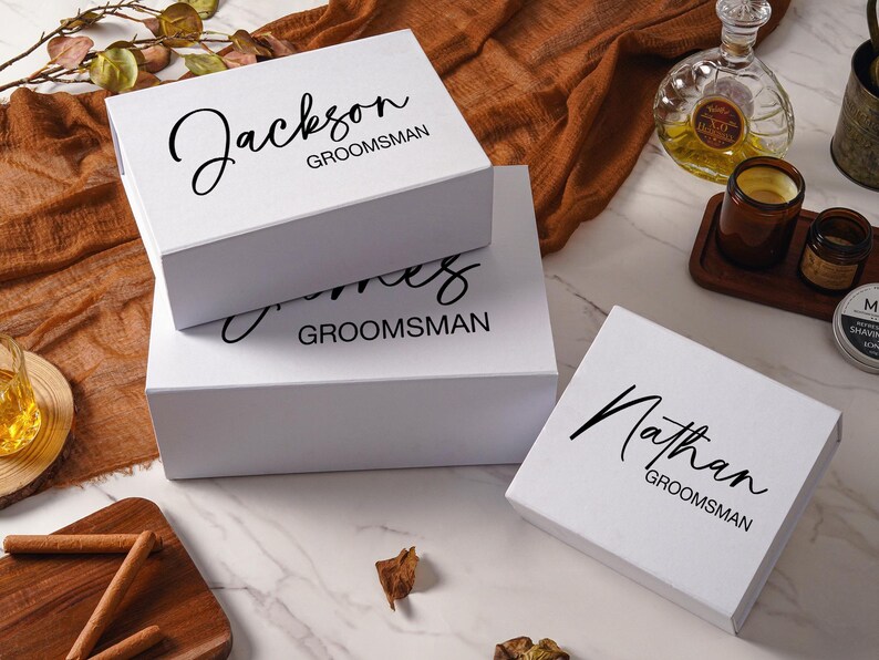 Peut inclure: Trois bo&icirc;tes cadeaux rectangulaires blanches avec du texte noir. La bo&icirc;te du haut porte l'inscription Jackson GROOMSMAN, la bo&icirc;te du milieu James GROOMSMAN, et la bo&icirc;te s&eacute;par&eacute;e &agrave; droite Nathan GROOMSMAN en m&eacute;langeant &eacute;criture cursive et b&acirc;ton.